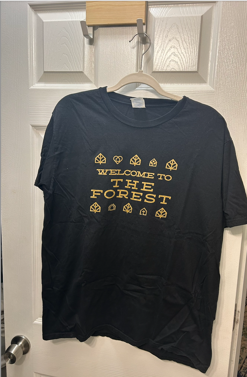 Black short-sleeve t-shirt with gold "Welcome to The Forest" graphic print and nature-themed icons.